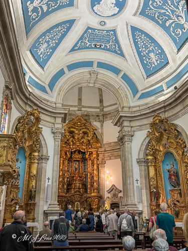 Interior view of a church with ornate golden altar and colorful ceiling, featuring visitors gathered for a service.