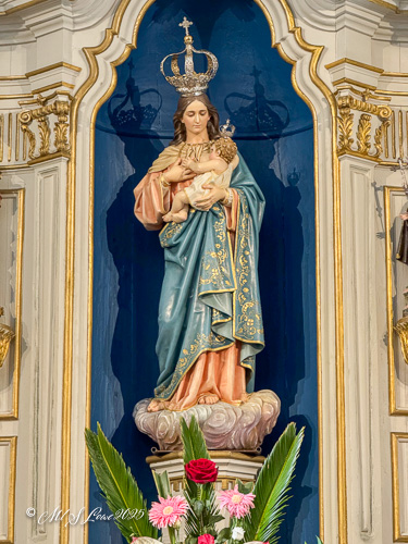 Statue of the Virgin Mary holding the infant Jesus, adorned with a crown, located inside a church, surrounded by floral arrangements.