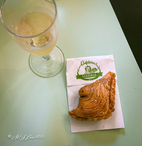 A glass of light-colored beverage alongside a flaky pastry on a napkin with a logo, set on a light-colored surface.