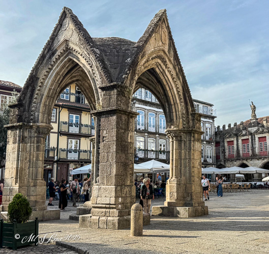 Historical stone arch in a bustling square, surrounded by people and traditional buildings.