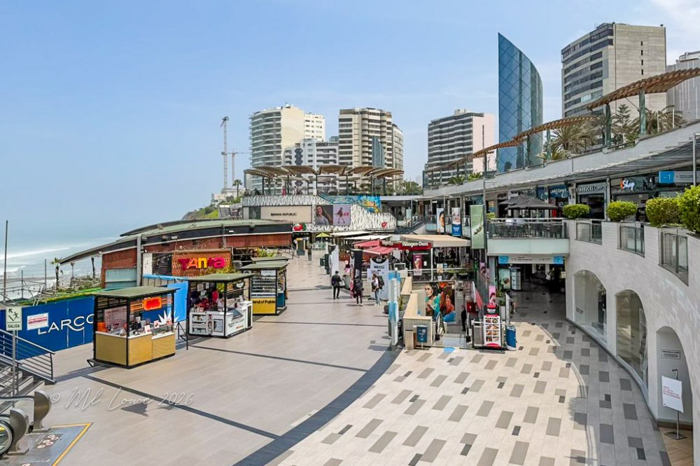 A view of a busy shopping promenade featuring various stalls, modern buildings, and palm trees, with the ocean in the background.