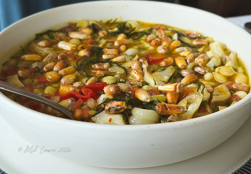 A bowl of vegetable stew featuring a mix of beans, corn, diced tomatoes, and leafy greens in a savory broth.