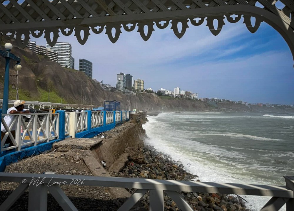 View of a coastal promenade with waves crashing against the shore, framed by an ornate structure. The skyline features modern buildings and a steep cliff in the background.