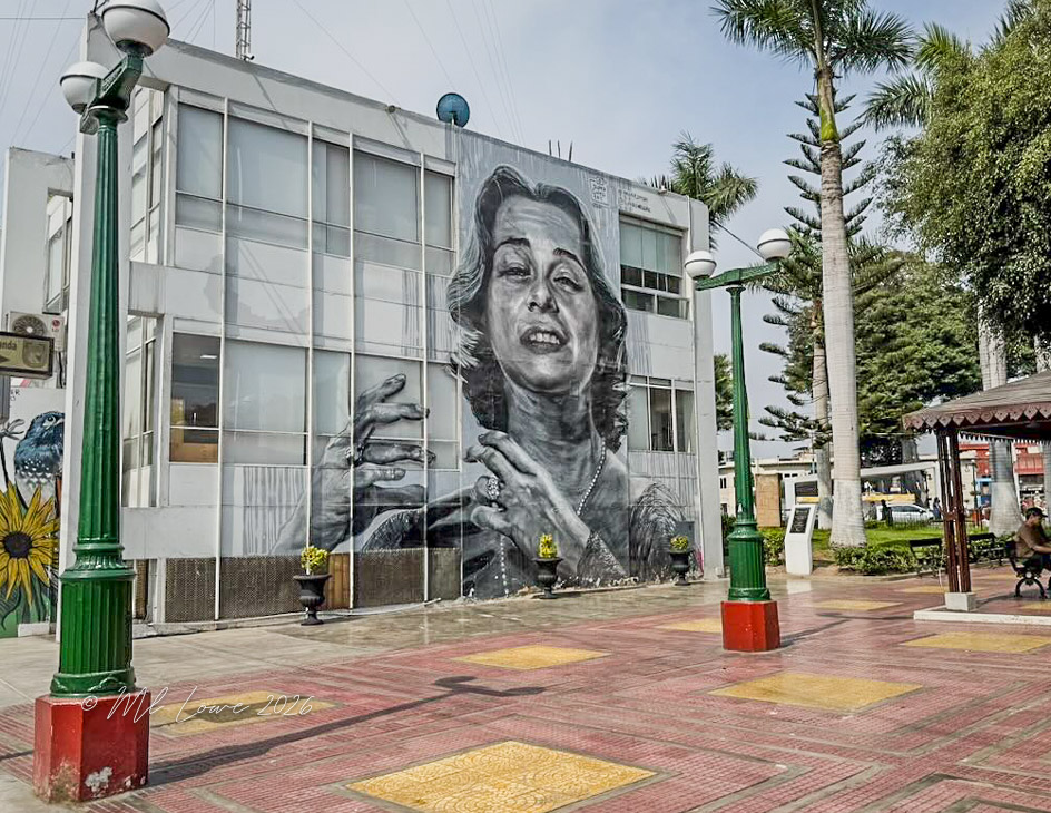A large mural of a woman with expressive hands is painted on the side of a building in a public square. Surrounding the area are decorative lampposts, palm trees, and benches.