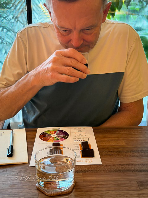 A man sitting at a wooden table, sipping from a small glass while looking at a tasting chart and notes, with a glass of water in front of him.