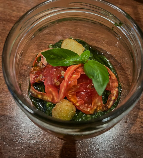 A small glass jar containing layers of green pesto, topped with sliced dried tomatoes, crispy croutons, and fresh basil leaves.