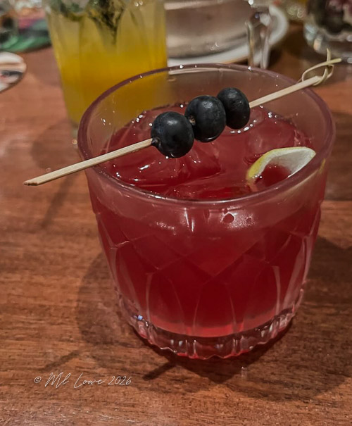 Close-up of a cocktail in a crystal tumbler, featuring a deep red color garnished with three blueberries and a slice of lime.
