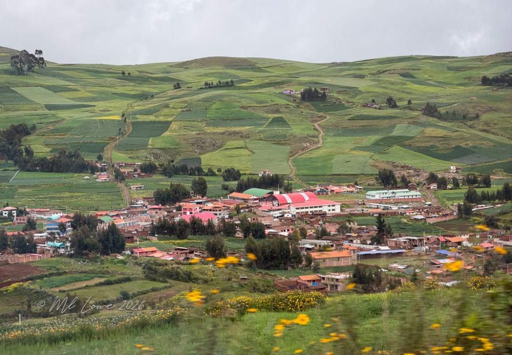 A panoramic view of a rural village surrounded by lush green fields and hills, with scattered houses and a prominent red-roofed building.