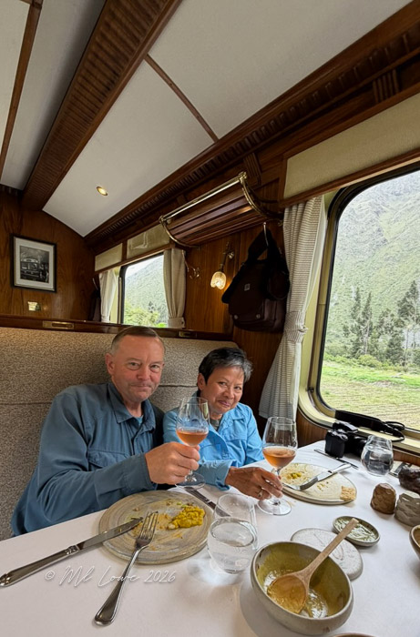 A couple enjoying a meal in a luxurious train dining car, toasting with glasses of pink drinks, surrounded by elegant wooden decor and scenic views from the windows.