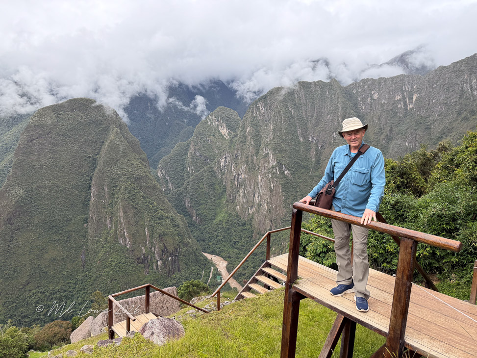 A man stands on a wooden platform overlooking the lush green mountains and valleys of Machu Picchu, with clouds hovering above.