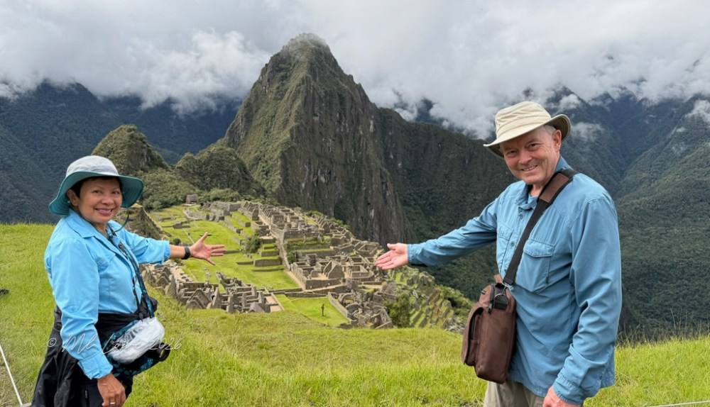 Two people smiling and pointing towards the ancient ruins of Machu Picchu, with mountains and cloudy skies in the background.