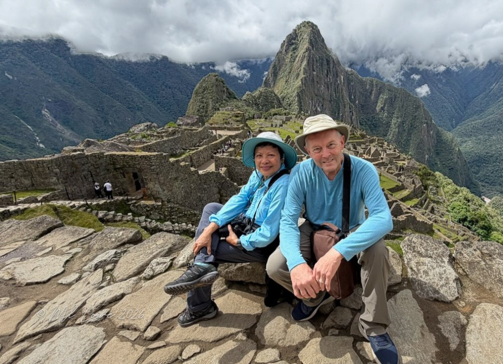 A man and a woman sitting on ancient stones at Machu Picchu, with lush green mountains and partly cloudy skies in the background.