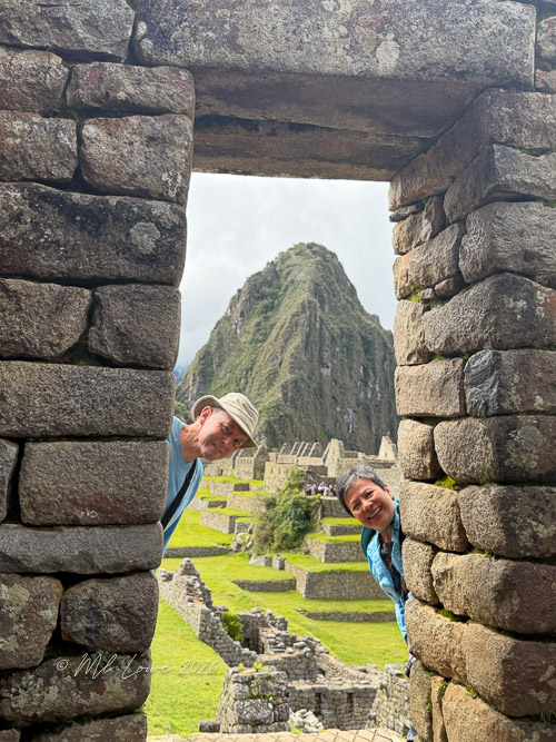 Two people peeking through a stone doorway at Machu Picchu, with a mountain and ancient structures visible in the background.