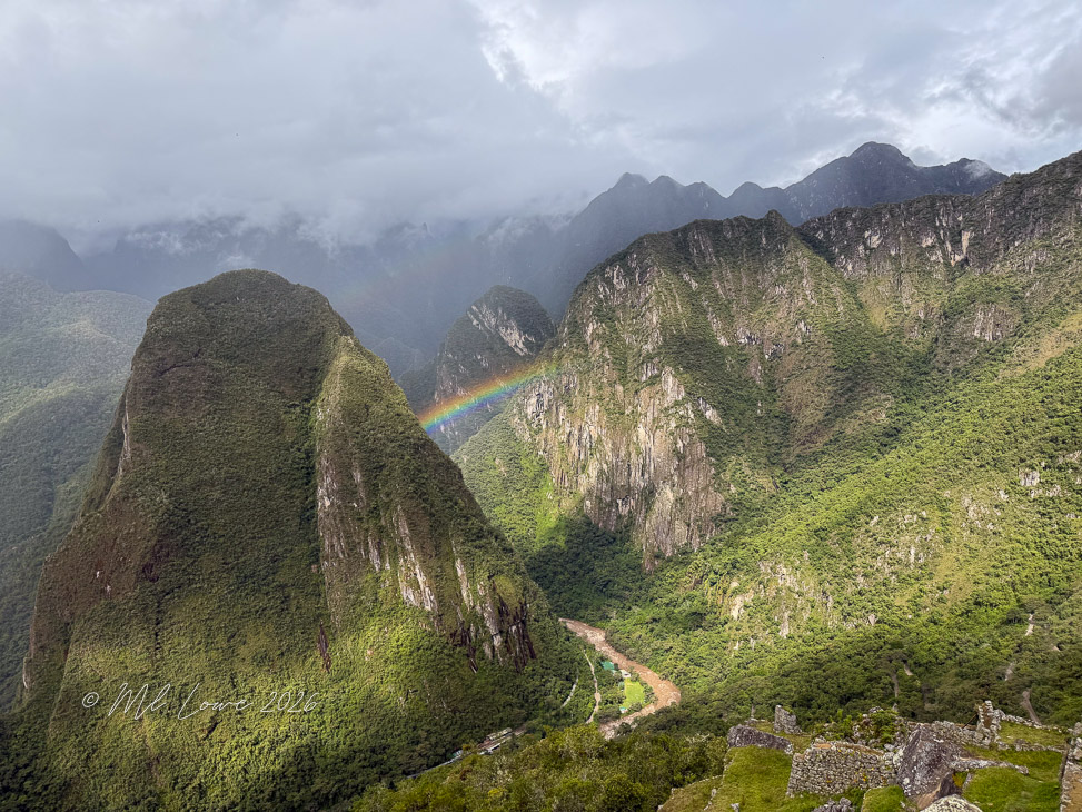 A scenic view of mountains and lush greenery with a rainbow arching through the sky, captured in a cloudy atmosphere.
