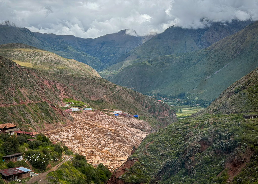 A panoramic view of the town of Maras with terraced salt pools in the foreground, surrounded by green hills and mountains under a cloudy sky.