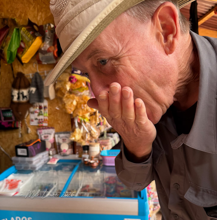 A close-up of a man wearing a hat, covering his mouth with his hand, appearing surprised or amused, in front of a colorful shop displaying snacks and goods.