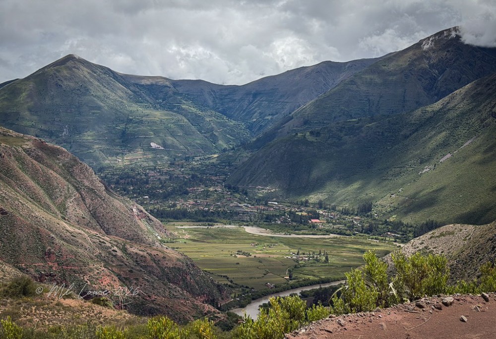 A panoramic view of a lush valley surrounded by steep mountains, with a river winding through the landscape and small settlements visible below under a cloudy sky.