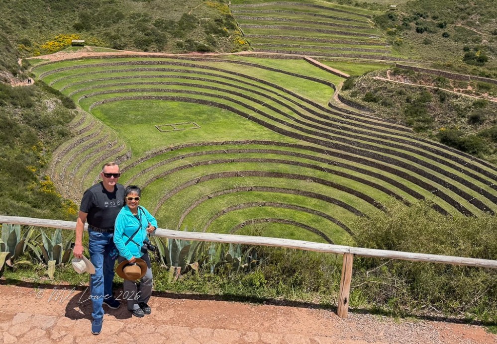 A couple stands in front of the Moray agricultural terraces, showcasing circular, stepped farming plots surrounded by lush greenery.