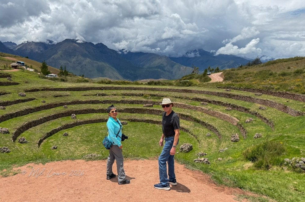 Two people walking in a circular agricultural terrace with mountains in the background, under a cloudy sky.