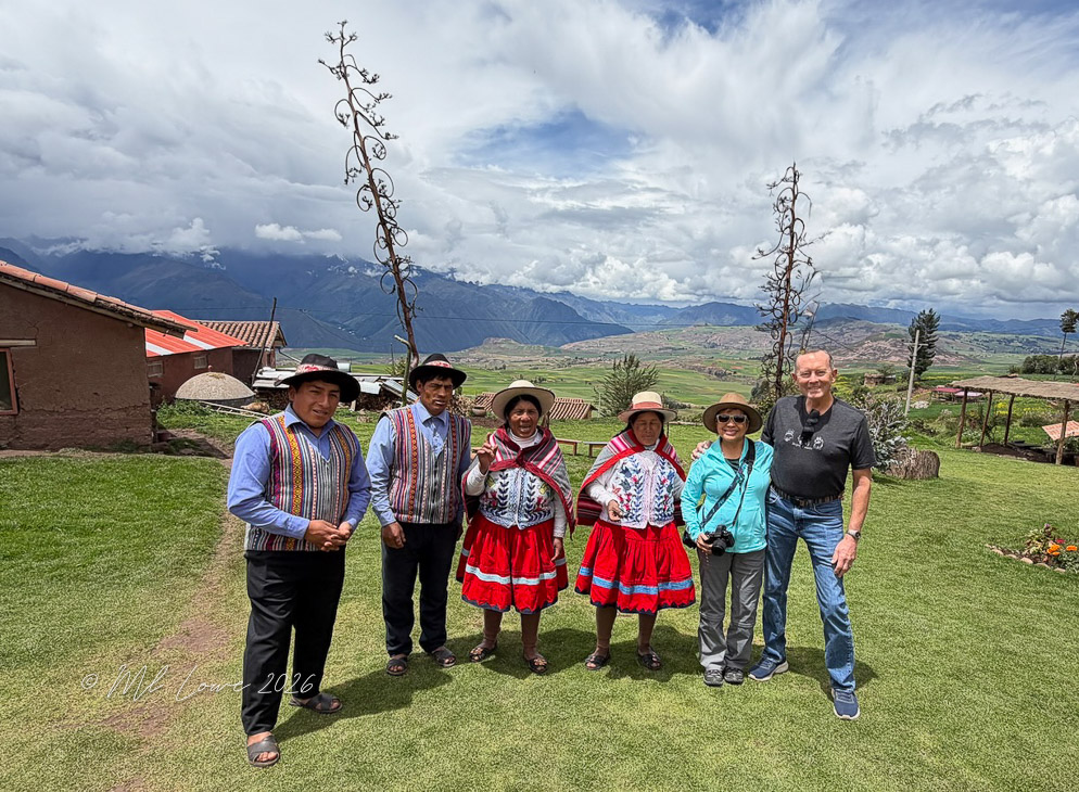 A group of six people, including two men in traditional Peruvian attire and four women in colorful dresses, stand on a grassy area with mountains in the background under a partly cloudy sky.