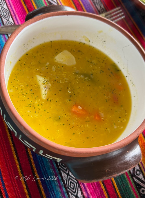 A bowl of yellow soup with vegetables and quinoa, placed on a colorful woven textile.