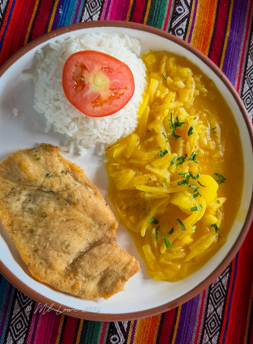 A plate of rice topped with a slice of tomato, served alongside sautéed onions and a piece of fried fish, on a colorful woven textile background.