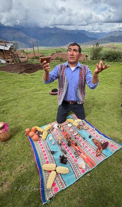 A man in traditional clothing kneels on a colorful blanket in a grassy field, holding a small clay pot in one hand while gesturing with the other. Various natural items like corn and herbs are arranged on the blanket, with mountains visible in the background under a cloudy sky.