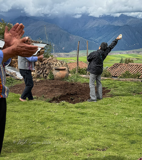 A group of people participating in a traditional ceremony outdoors, with men clapping and one man raising an item towards the sky amid a mountainous backdrop.