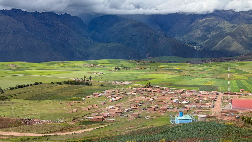 A panoramic view of a rural area with a small village surrounded by lush green fields and distant mountains under a cloudy sky.