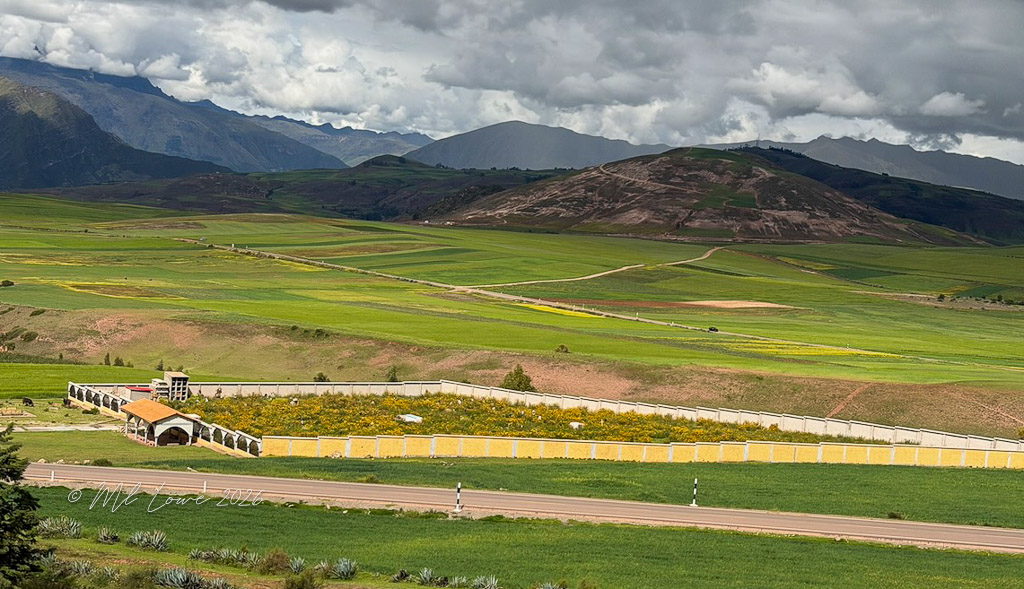 A scenic view of lush green fields with a mountain range in the background, featuring a neatly enclosed garden area with colorful flowers and a structure. The sky is partly cloudy, enhancing the natural beauty of the landscape.