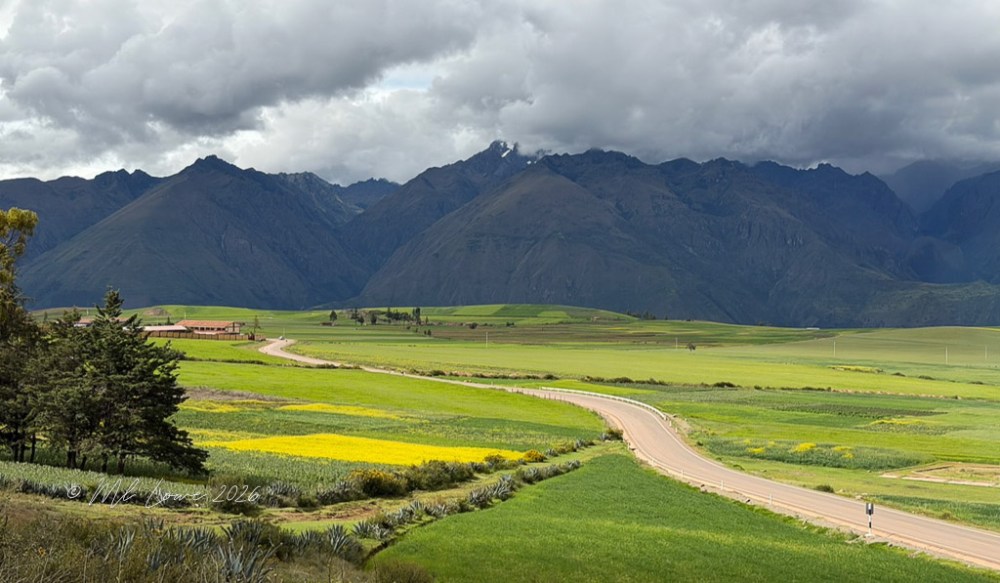 Panoramic landscape view of rolling green fields with a winding road, surrounded by majestic mountains under a cloudy sky.