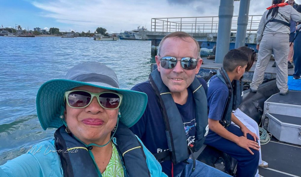 Two people smiling for a selfie on a boat, wearing life jackets, with a waterfront and boats in the background.