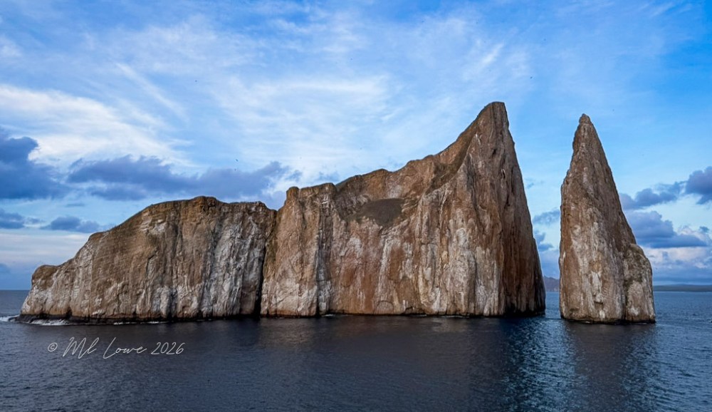 Two tall rock formations rising from the ocean under a cloudy blue sky.