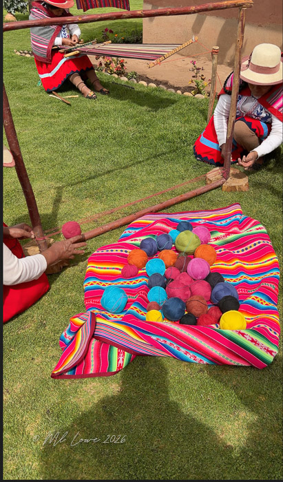 Two women in traditional Peruvian clothing work with colorful yarn on a grassy lawn. One woman is sitting at a weaving frame, while the other is arranging various vibrant yarn balls on a multicolored striped blanket.