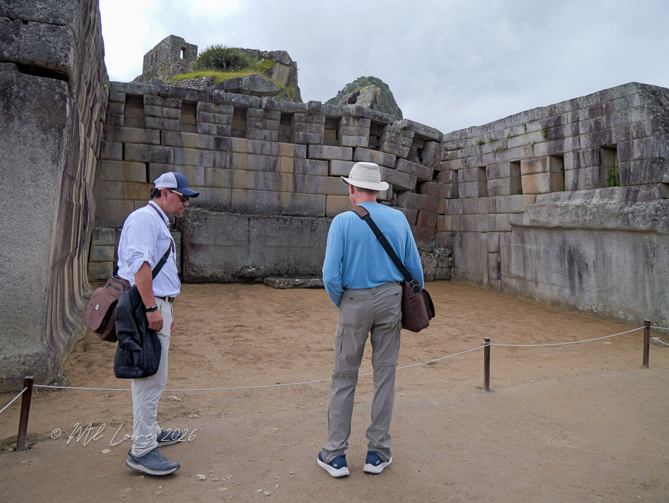 Two men exploring an archaeological site with large stone walls and a sandy floor, set against a cloudy sky.