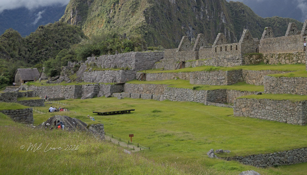 Ruins of ancient stone structures at Machu Picchu, surrounded by lush greenery and steep mountains, with visitors exploring the site.