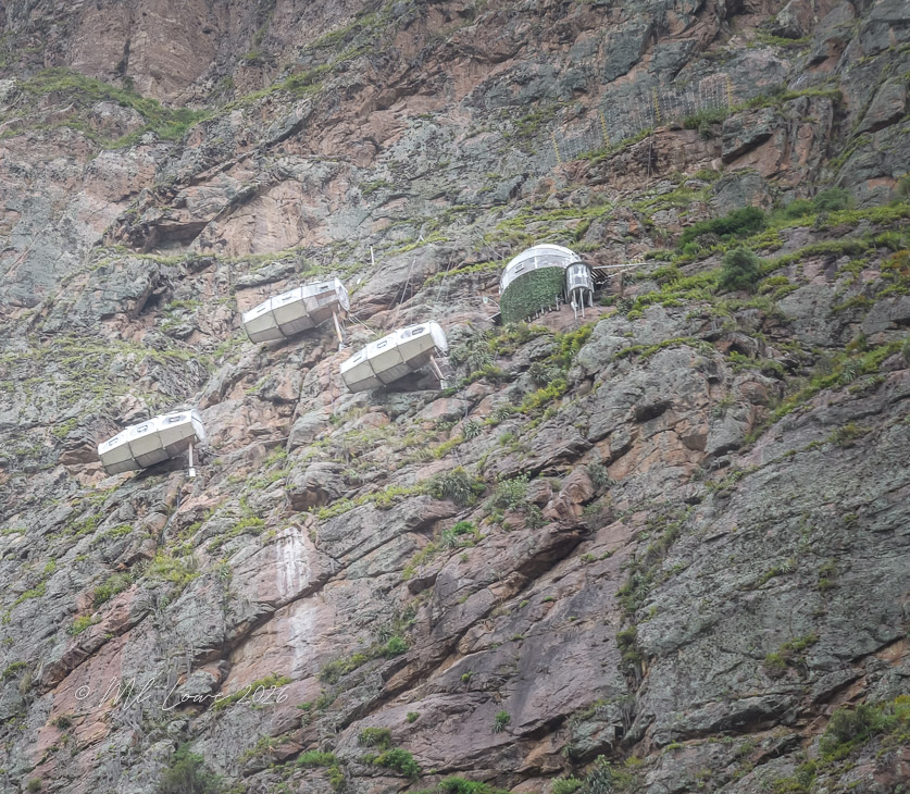 Three unique glass cabins perched on a rocky mountainside, surrounded by green vegetation.