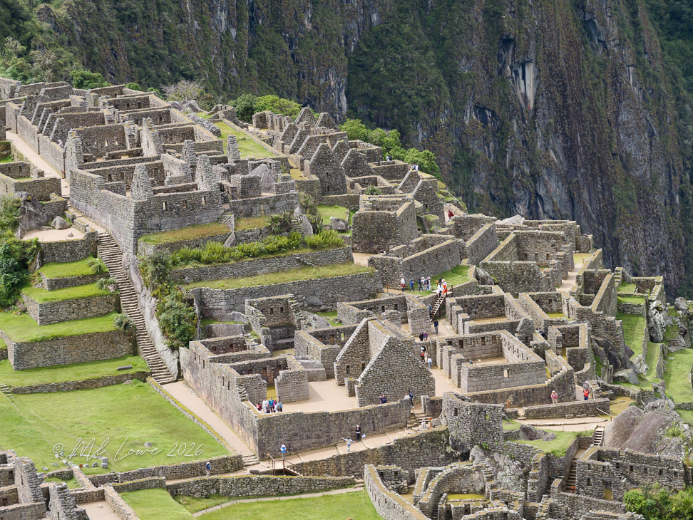 Aerial view of ancient stone structures at Machu Picchu, surrounded by lush greenery and steep cliffs. Visitors explore the ruins.