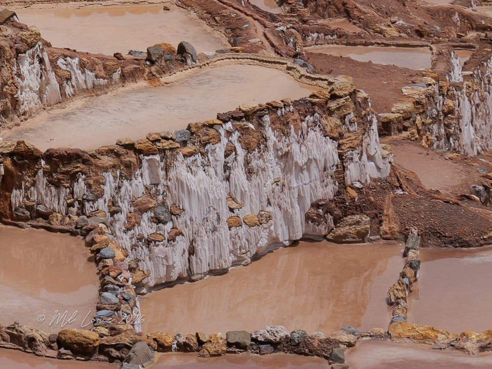 Aerial view of salt flats with terraced pools and rocky edges, displaying layers of salt deposits and muddy water.