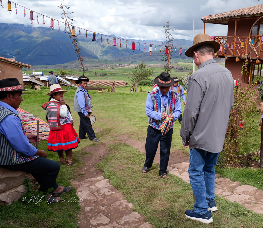A group of individuals in traditional attire interacting on a path in a rural setting, with mountains and colorful decorations in the background.