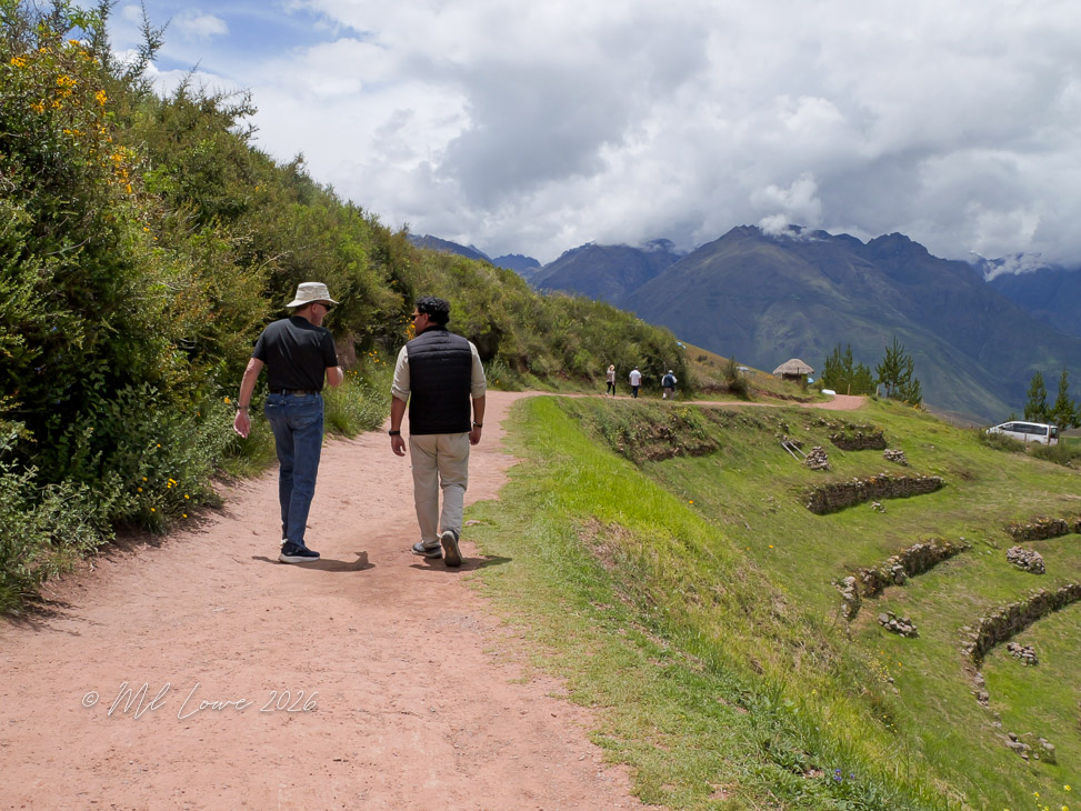 Two men walking along a dirt path in a hilly landscape with green vegetation and mountains in the background. One man wears a hat and a black shirt, while the other wears a lightweight vest. In the distance, additional people are walking, and there are stone agricultural terraces on the hillside.