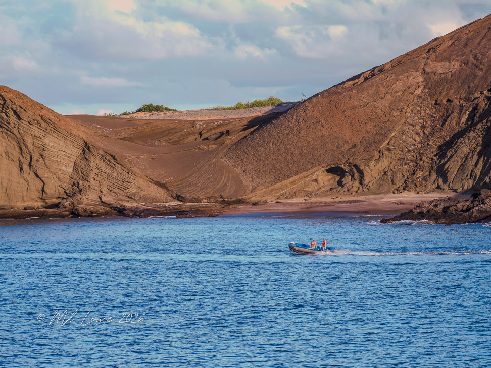 A small boat with two people navigating through blue water, with rugged brown hills and sandy beaches in the background under a partly cloudy sky.