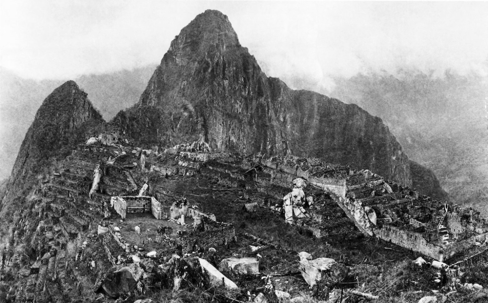 Black and white image of Machu Picchu ruins nestled in the Andes mountains, with steep cliffs and foggy background.