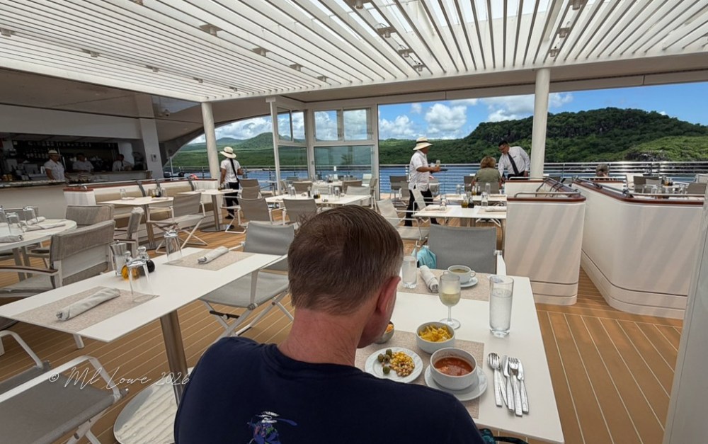 A man seated at a table in a modern outdoor dining area, enjoying a meal with various dishes, while staff in uniforms serve other guests in the background. The scene features a view of greenery and water outside.
