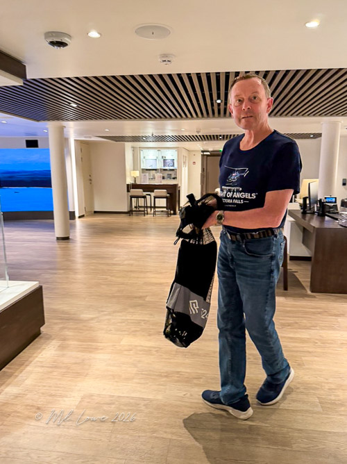 A man holding a black and gray bag, wearing a blue t-shirt and jeans, standing in a well-lit indoor space with a modern design.