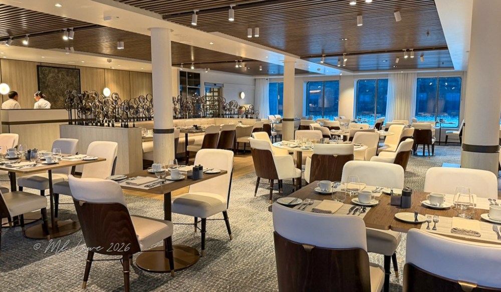 A modern restaurant interior featuring elegantly set tables with white and wooden chairs, large windows revealing a scenic view, and chefs visible in the background.
