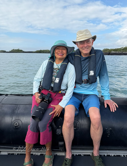 A woman and a man sitting on the edge of a small boat in a body of water, both wearing life jackets and casual clothing. The woman holds a camera, and they appear to be enjoying a day out on the water.