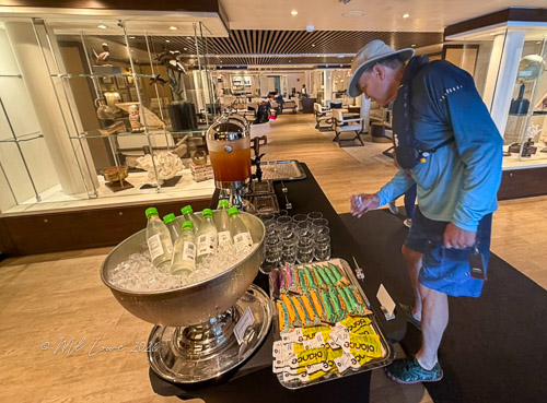 A man with a backpack stands by a beverage station in a hotel lobby, selecting drinks from a table with ice, water bottles, and colorful snacks.