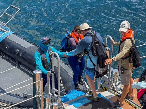Two people boarding a small boat from a dock, while another person assists them. The scene includes blue water and a small inflatable boat.