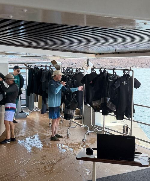 A group of people on a boat preparing for diving, with wetsuits hanging on a rack in the background and a laptop on a table.